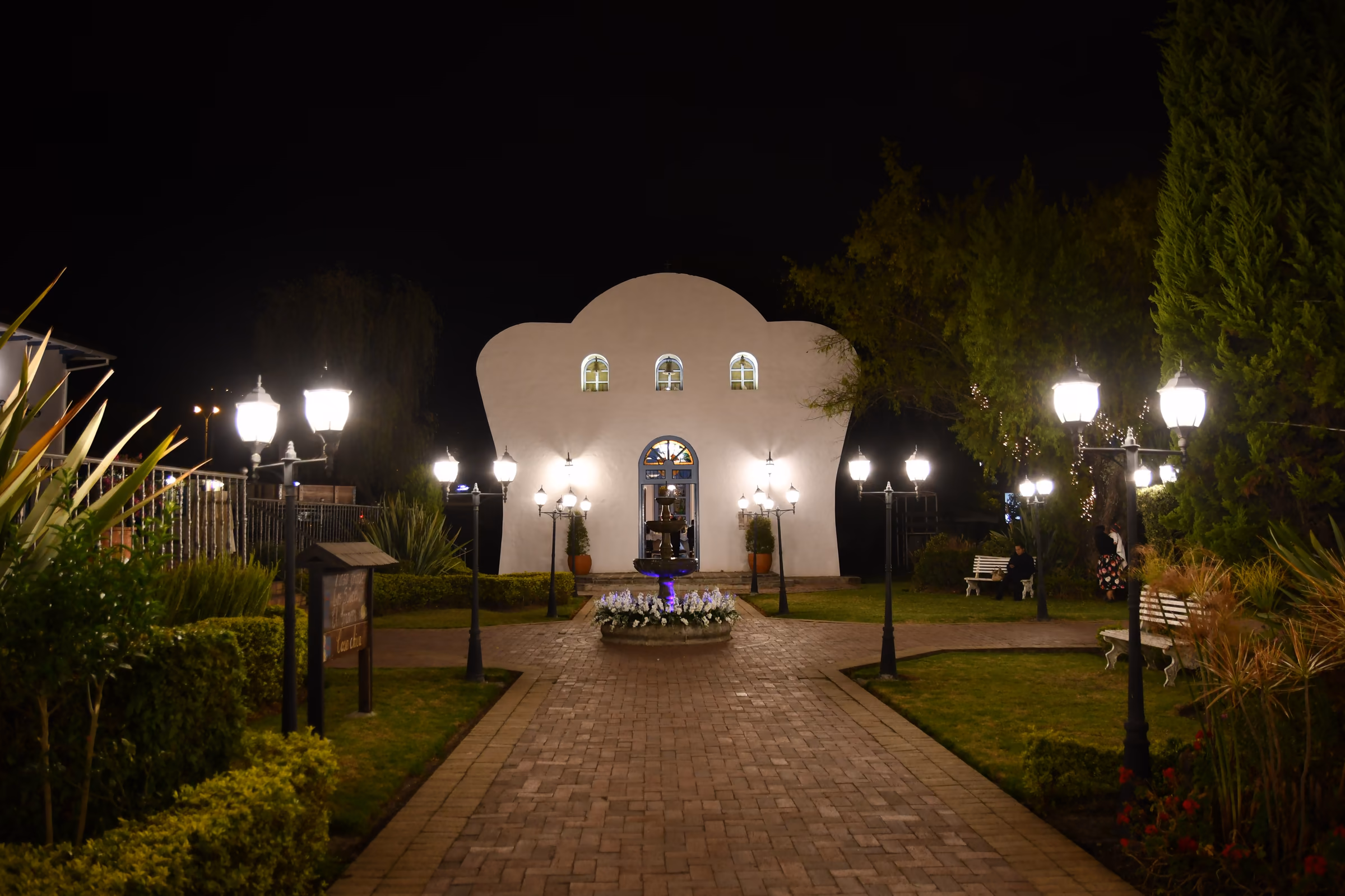 La capilla iluminada de noche al final del camino de ladrillo, entre faroles y jardineras.
