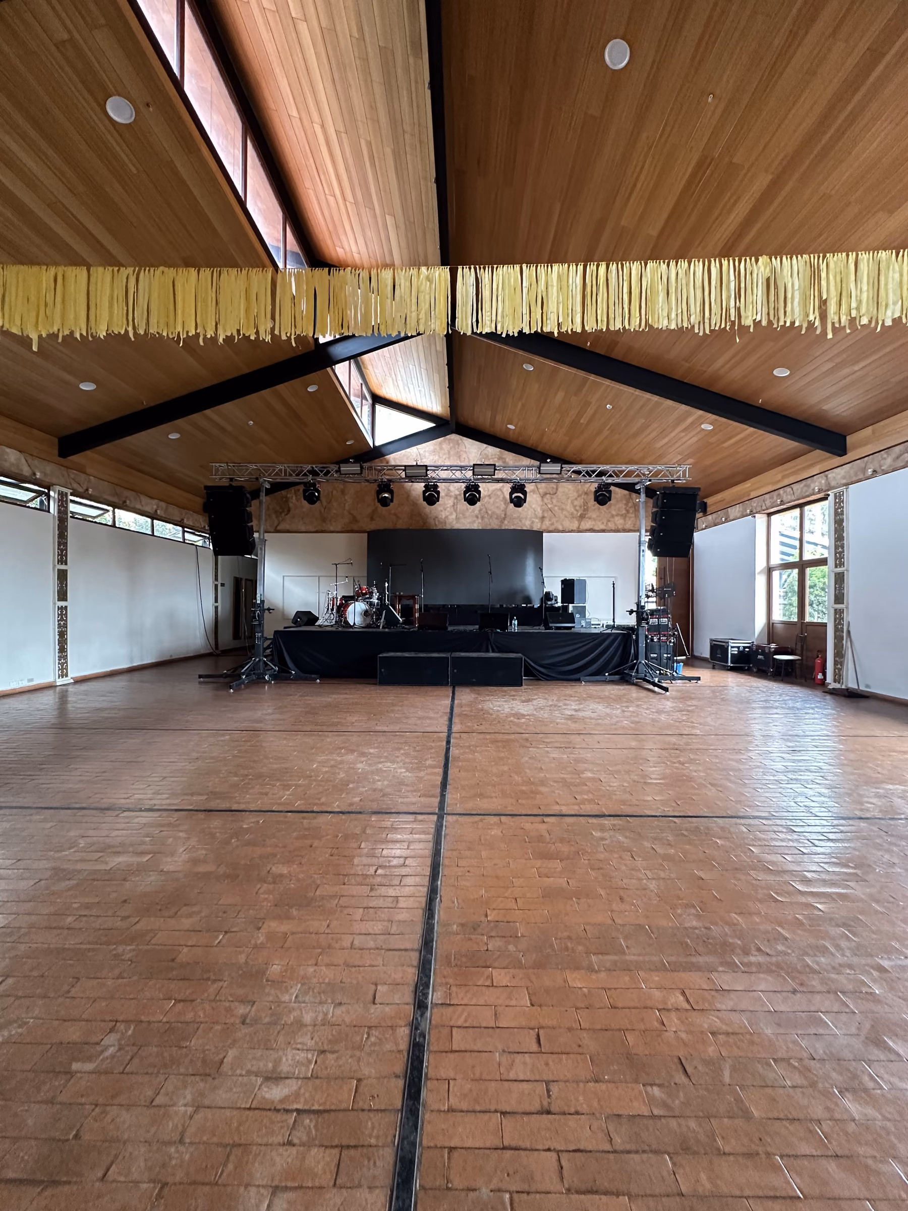 El salón vacío con tarima, torre de luces y equipo de sonido montados para un concierto, bajo el techo de madera a dos aguas.