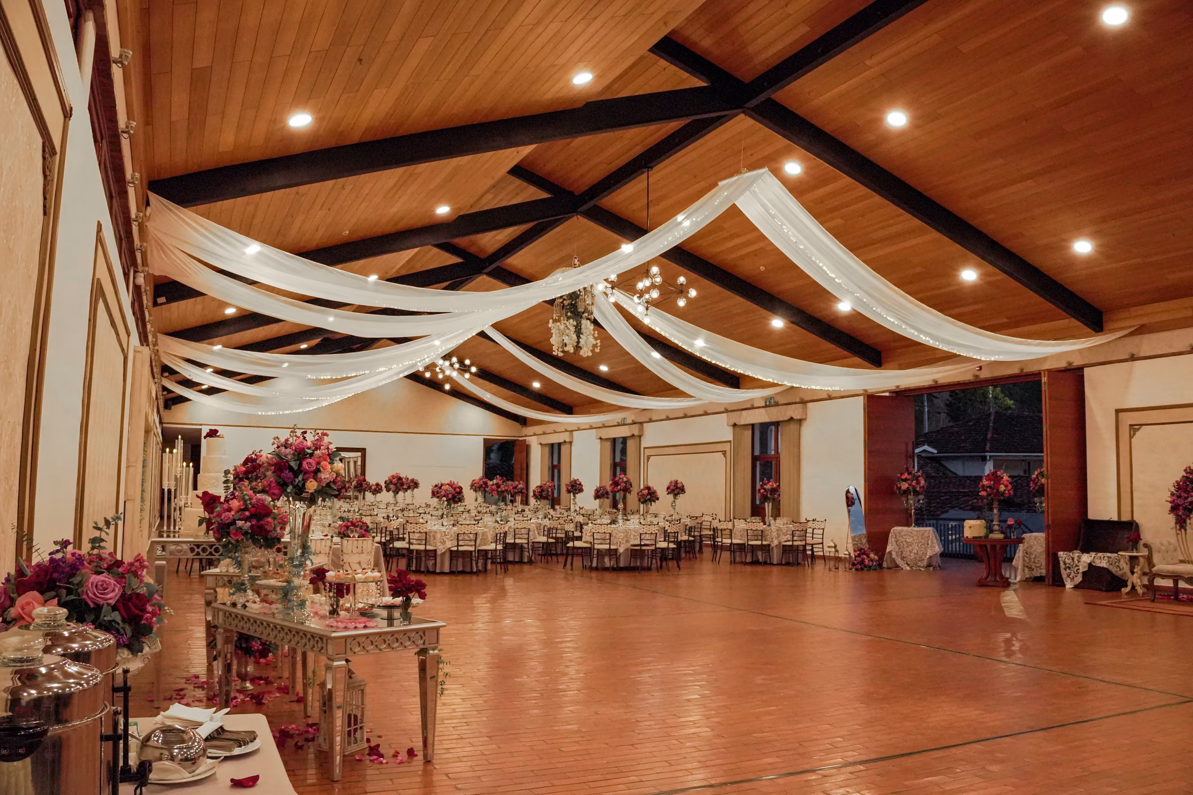 Salon Los Sauces montado para una boda de noche, con telas blancas cayendo desde el techo de madera y centros de flores fucsia sobre las mesas.